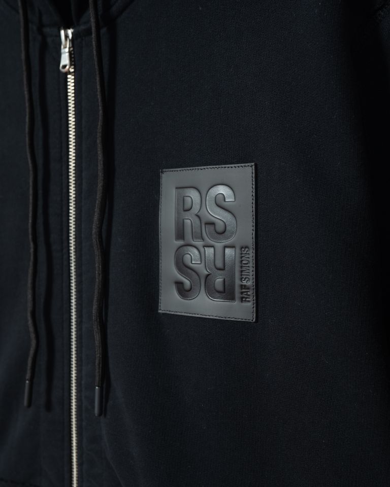 RAF SIMONSのジップアップフーディは、コットン100%の素材を使用しており、裏毛パイルの着心地の良さが特徴です。両肘にはRSハンドサインのグラフィックが施され、ユニークでアイコニックなデザインとなっています。また、RSのレザーパッチが付いており、ブランドの高級感を演出しています。 生地切り替え部分は切りっぱなしの仕上げとなっており、こだわりを感じるディテールに仕上がっています。