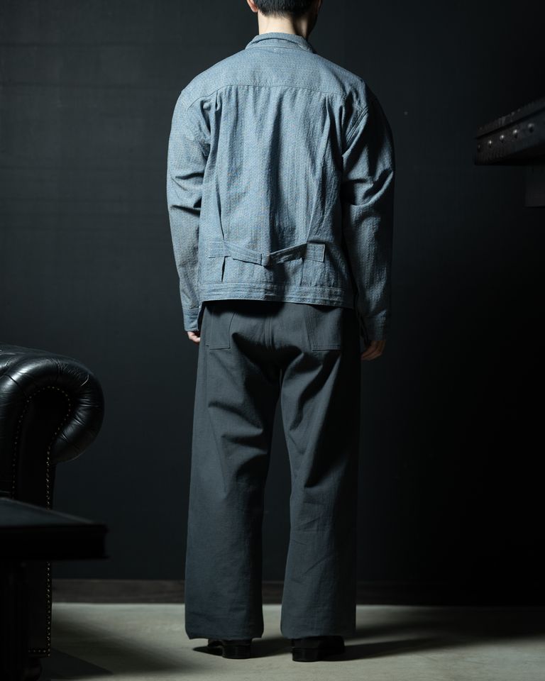 後ろ姿全体。肩傾斜のない水平な肩線が、背面を広くフラットに見せる。腰のバックルがシルエットのウエスト位置を視覚的に示し、ゆったりとした中にも構造的な美しさを持つ。