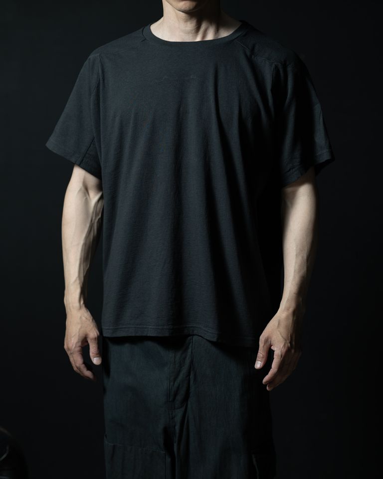 ドルマン調の袖付けにより肩まわりに余裕があり、体に沿わず少し浮くように落ちるシルエットが確認できる。首まわりのリブは細く、ネックラインが大きめに開いているため、すっきりと清潔感のある印象。暗めのライティングでチャコールブラックの色の深みがよく出ている。