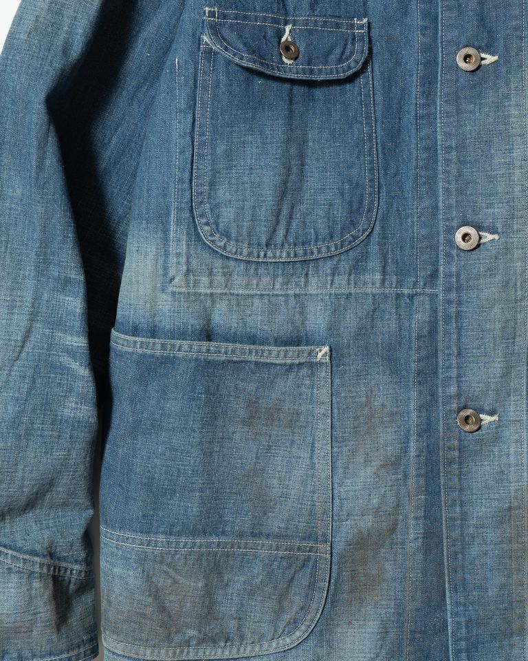 左前身頃のポケット詳細。フラップ付き胸ポケットと大容量のローポケットが機能的に配置され、鉄製ボタンの素材感と縫製のディテールが間近で確認できる。オイル汚れを模したダメージが生地の表情に奥行きを加えている。