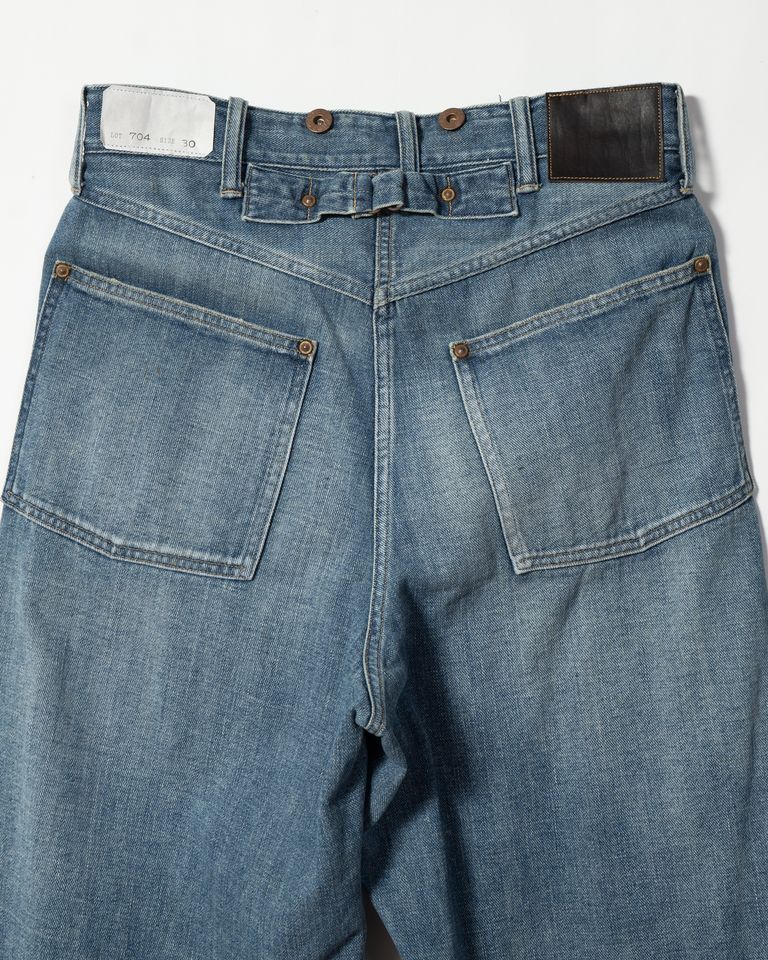 Taiga Takahashi のアイコニックアイテムでもある Lot.704 Demin Trousers c.1920s にダメージ加工を施したジーンズ。通常のダメージ加工に加え。塩縮加工を施すことで、生から穿いたような、シワ、ヒゲ等のユーズド感を実現した。錆びて朽ちたボタンとリベット、バックルが存在感を作り出している。