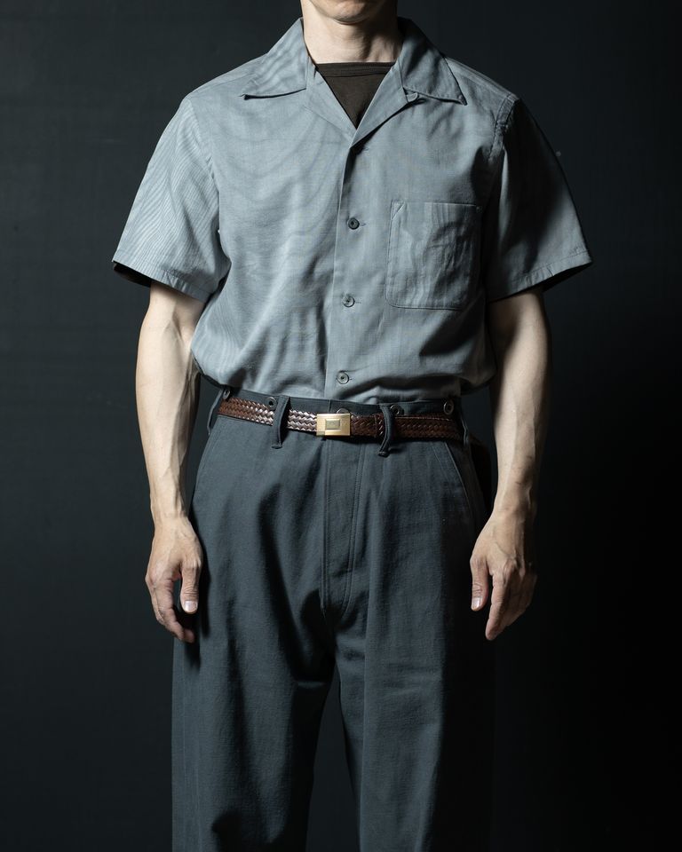 タックインスタイルの正面カット。編み込みレザーベルトを合わせることでウエストラインが生まれ、シャツの丈感の汎用性を示す一枚。絣調素材の微細な縦縞と胸ポケットのディテールがよく見て取れる。