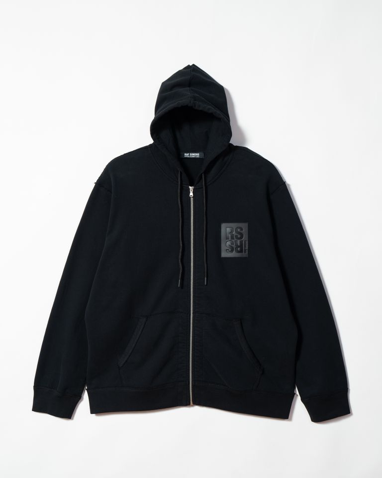 RAF SIMONSのジップアップフーディは、コットン100%の素材を使用しており、裏毛パイルの着心地の良さが特徴です。両肘にはRSハンドサインのグラフィックが施され、ユニークでアイコニックなデザインとなっています。また、RSのレザーパッチが付いており、ブランドの高級感を演出しています。 生地切り替え部分は切りっぱなしの仕上げとなっており、こだわりを感じるディテールに仕上がっています。