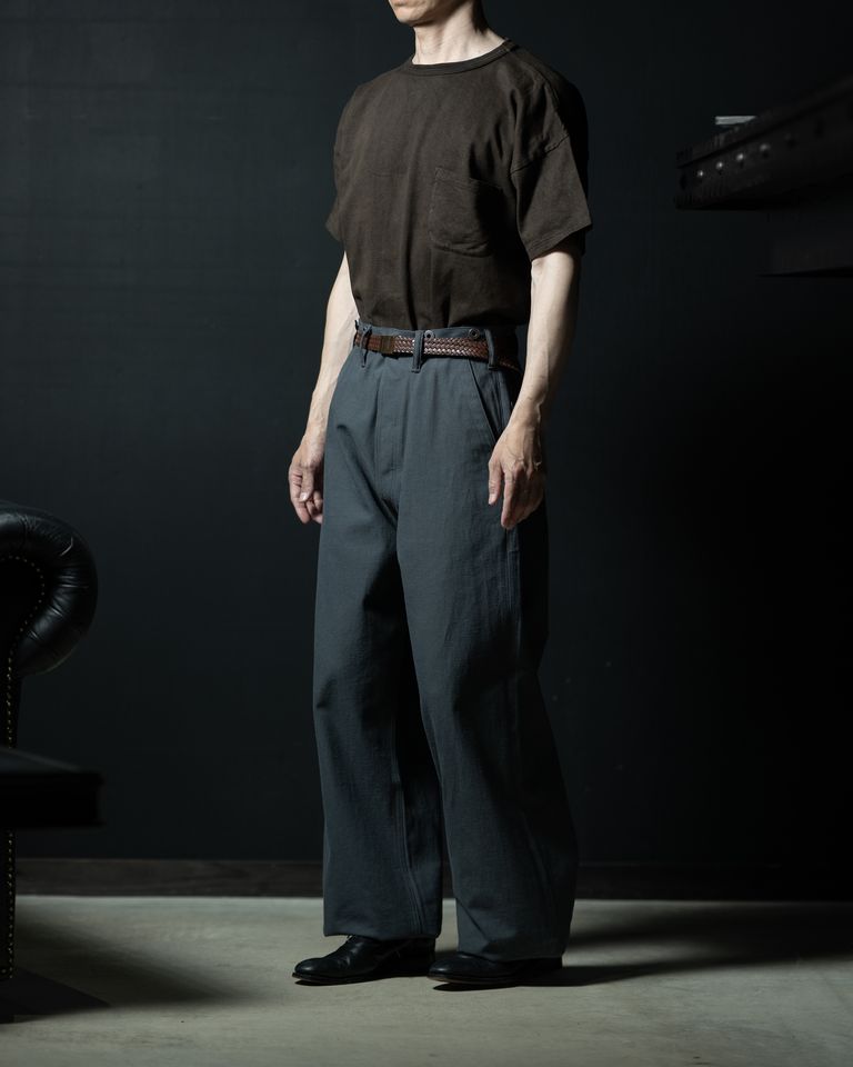 サイドからの全身タックインコーデ。ブラウンのTシャツとグレーのワイドパンツが柔らかいコントラストを生み出し、黒いレザーシューズが全体を引き締める。チェスターフィールドソファが背景に佇む。