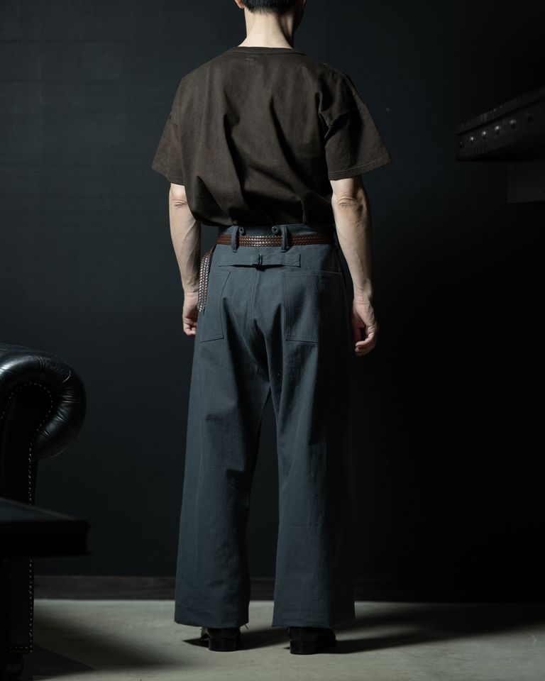 ベルトスタイリングの後ろ全身カット。グレーワイドパンツのバックポケットとシンプルなベルトループが、泥染めブラウンの無骨さと品よく調和する。チェスターフィールドソファが空間全体に風格を与える。