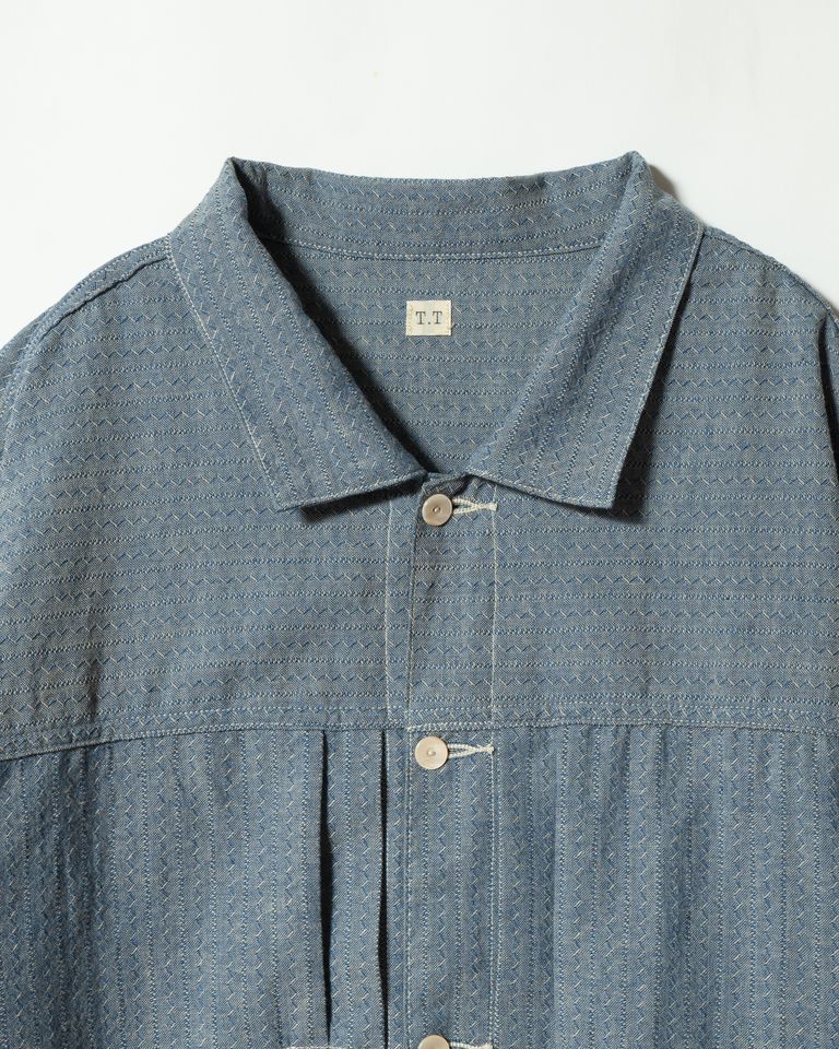 衿元のクローズアップ。「T.T」のネームタグとともに、貝ボタンを糸で結ぶオリジナルのボタン付け仕様が確認できる。衿の丸みと開き具合が、シャツとしての顔を持つこのジャケットの表情を決定づけている。