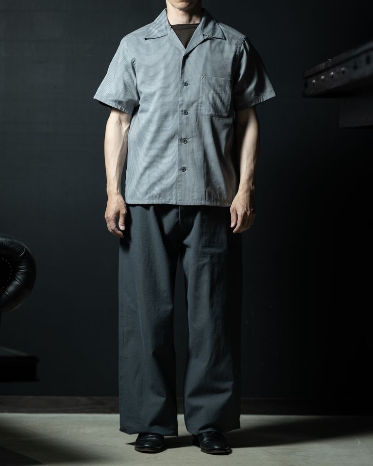 ワイドパンツとのセットアップ風コーディネート。シャツのグレイッシュなブルーとパンツのダークグレーが同系色でまとまり、モノトーンの落ち着いた佇まいを見せる。裾をアウトにしたノータックインスタイルで、全身にゆとりのあるシルエットを表現。