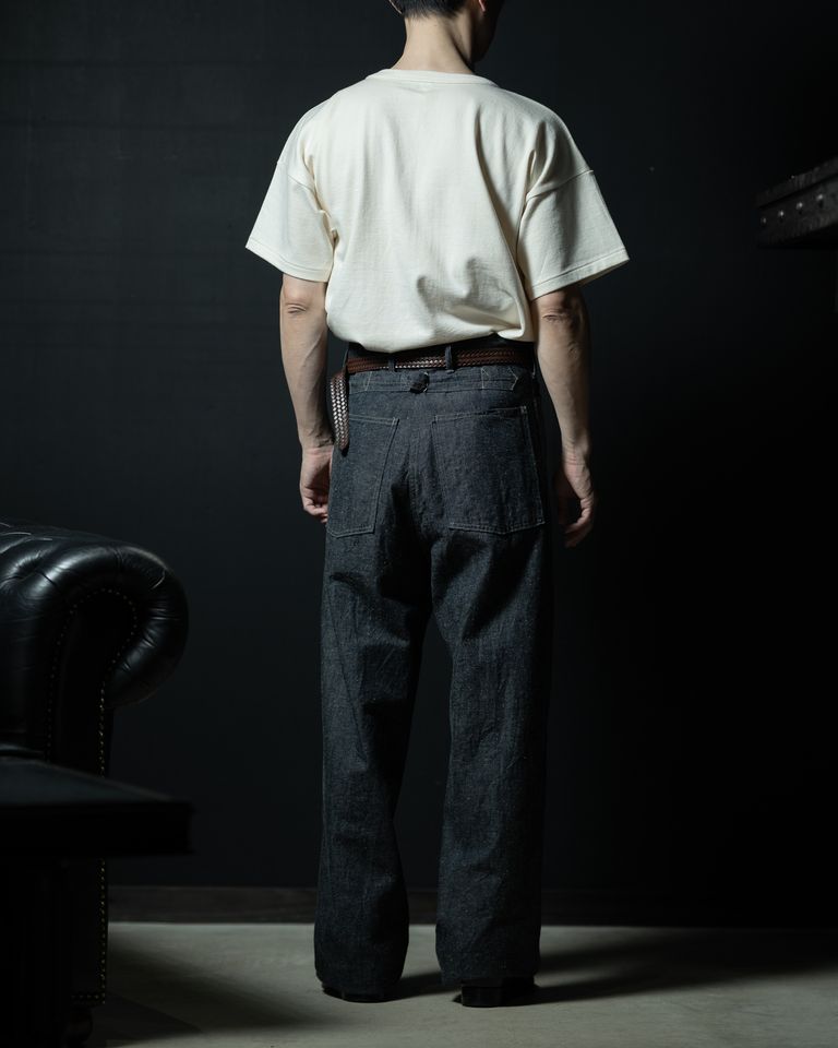ベルトスタイリングの後ろ全身カット。ワイドデニムのバックポケットのステッチディテールと、Tシャツの生地感が互いを引き立て合う。チェスターフィールドソファが空間全体に格を与える。