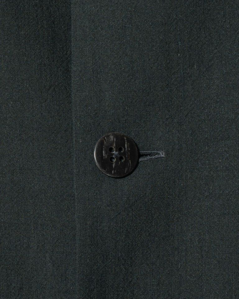 リネンとシルクの交織が作る縦の節感と、黒蝶貝ボタンの質感が同一フレームに収まる。ボタン穴はループ式ではなく縫い穴タイプ。生地の織り目を見ると、リネンの粗い糸の間にシルクの光沢がのっているのが分かる。触れる前から伝わる素材の密度感。